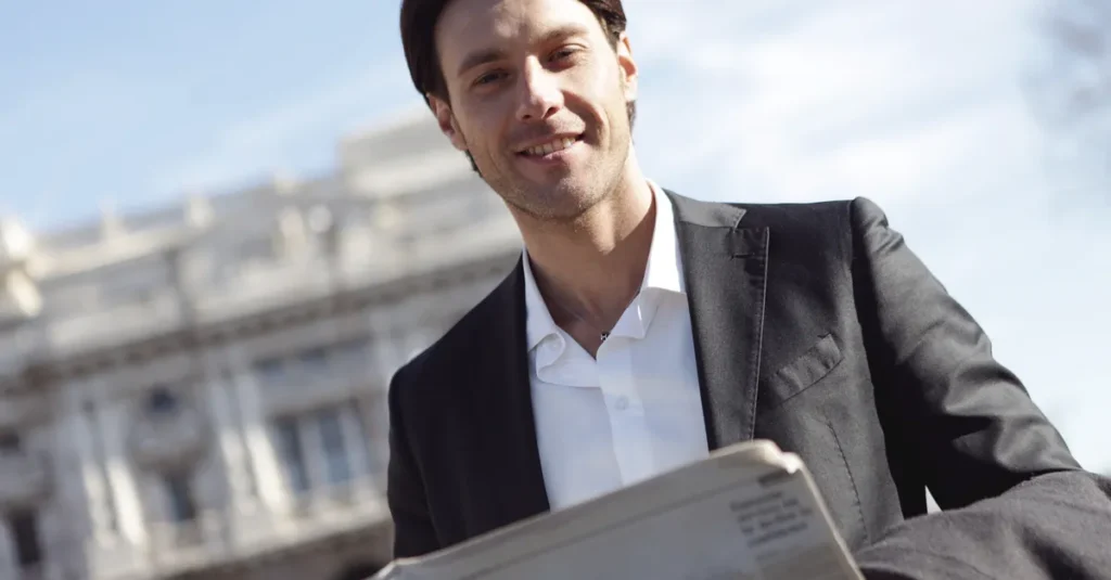 Smiling businessman in suit reads newspaper outside on a sunny day.