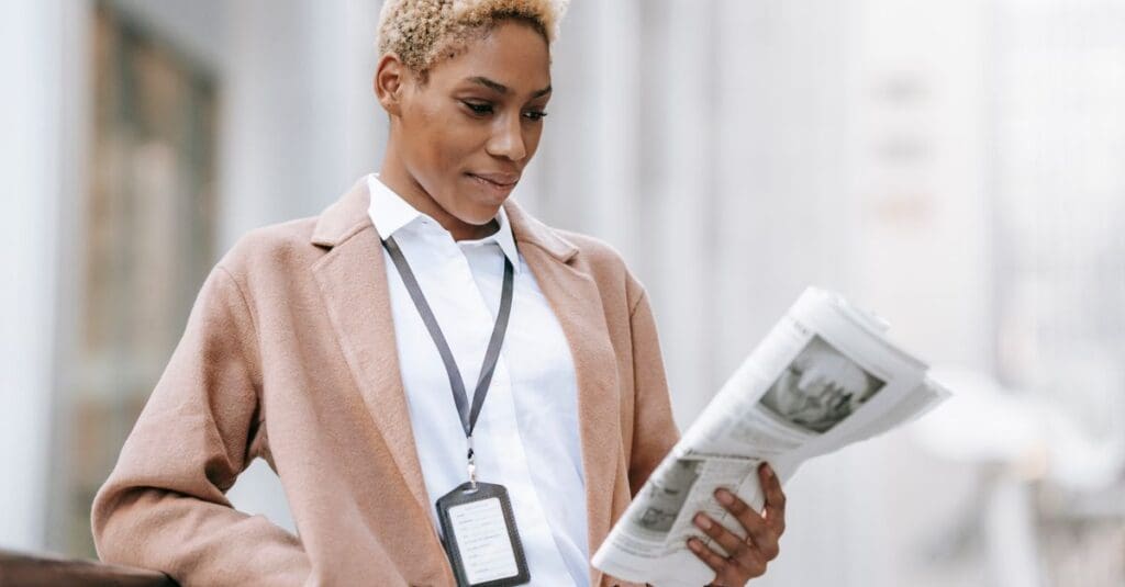 Serious young African American female entrepreneur with badge in elegant brown coat standing near railing and reading newspaper