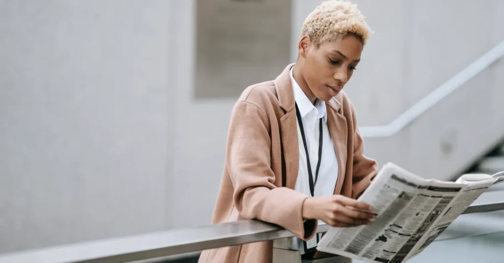 Serious young African American businesswoman in formal clothes leaning on business center building railing and reading article in newspaper in daylight