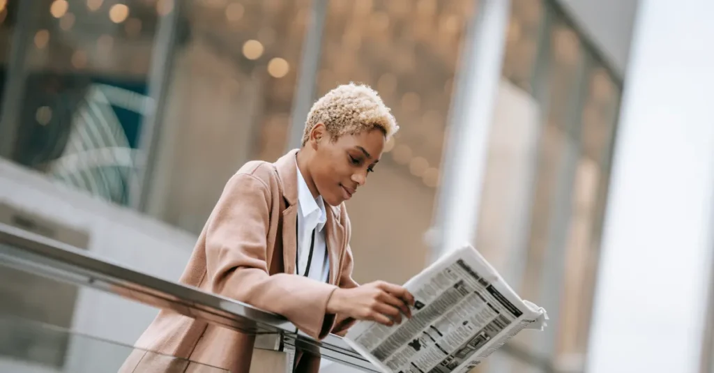 Young attractive African American businesswoman in formal clothes reading newspaper and leaning on glass railing while standing outside contemporary office building